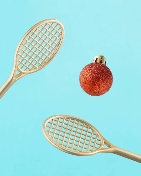 New Year sport or recreation concept with Golden tennis rackets and red decorative Christmas tree ball with glitters on isolated pastel blue background. Minimal creative abstract aesthetic Xmas card.