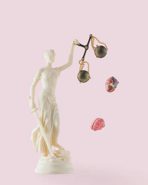 Heart and brain fall out of Lady Justice scales on isolated pastel pink background. Punishment unbalanced between emotions and rationality. Critical concept of law, anger, injustice. Human head idea.