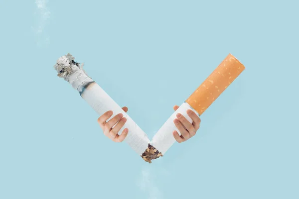 V-shaped victory symbol made with broken cigarette held by man\'s hands on isolated pastel blue background. Minimal creative abstract surrealist poster of  World no tobacco day. Quit smoking card.