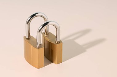 Parlak metalik prangalı pirinç asma kilitler, gölgeli bej arka planda izole edilmiş, güvenlik ve korumayı simgeliyor. Asgari güvenlik, birlik veya sağlam ilişki kavramı.