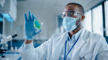 Yüz maskesi takan ve Petri Çanağına Genetik olarak modifiye edilmiş Kimyasallarla bakan siyah erkek bilim adamı. Modern Laboratuvarda Teknolojik Ekipmanlarla Çalışan Mikrobiyolog.