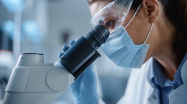 Mikroskop 'a bakan Maskeli ve Gözlüklü Kadın Bilimadamının Makro Yakın Çekimi. Modern Laboratuvarda Teknolojik Ekipmanlarla Molekül Örnekleri üzerinde çalışan Kadın Mikrobiyolog.