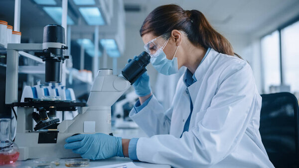 Female Scientist in Face Mask and Glasses Looking a Petri Dish with Genetically Modified Sample Chemicals Under a Microscope. Microbiologist Working in Modern Laboratory with Technological Equipment.