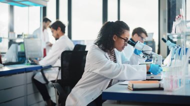 Tıp Bilimi Laboratuvarı: Mikroskop Altında İnceleme Yapan Güzel Siyah Bilim Adamı Test Örnekleri Yapıyor. Gelişmiş Teknoloji Malzemeleri Kullanan Çeşitli Genç Uzmanlar Ekibi.