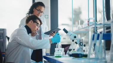 Modern Tıbbi Araştırma Laboratuvarı: Sıvı Kimyasalları Pipette, Dijital Tablet ve Konuşarak Karıştıran İki Genç Bilim Adamının Portresi. Gelişmiş Laboratuvar 'da çok ırklı uzmanlardan oluşan çeşitli bir ekip çalışıyor.