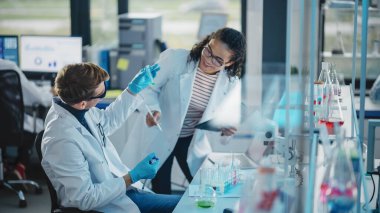 Modern Tıbbi Araştırma Laboratuvarı: Sıvı Kimyasalları Karıştıran İki Genç Bilim Adamının Portresi, Sonuçları Denetleme. Dijital tablet, konuşma. Gelişmiş Laboratuvar 'da çok ırklı uzmanlardan oluşan çeşitli bir ekip çalışıyor.