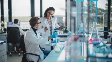 Modern Tıbbi Araştırma Laboratuvarı: Sıvı Kimyasalları Pipette, Dijital Tablet ve Konuşarak Karıştıran İki Genç Bilim Adamının Portresi. Gelişmiş Laboratuvar 'da çok ırklı uzmanlardan oluşan çeşitli bir ekip çalışıyor.