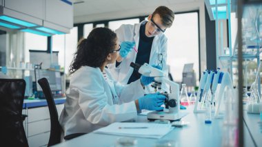 Modern Tıbbi Araştırma Laboratuvarı: İki genç bilim adamının Portresi Mikroskop, Örnek Analiz Yapıyor, Konuşuyor. Gelişmiş Laboratuvar 'da çok ırklı uzmanlardan oluşan çeşitli bir ekip çalışıyor.