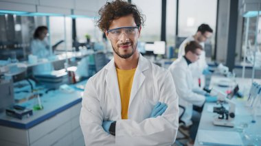 Beyaz önlüklü, güvenlik gözlüklü, çapraz kollu, kameraya bakan yakışıklı Latin bilim adamı. Genç Biyoloji Teknolojisi Araştırma Uzmanı. Orta Resim Çekimi