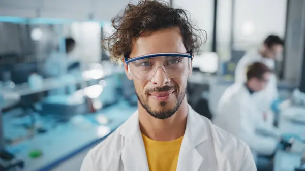 Medical Science Laboratory: Handsome Young Latin Scientist Wearing White Coat and Safety Glasses Smiles Looking at Camera. Young Bio Technology Research Specialist. Medium Closeup Portrait Shot