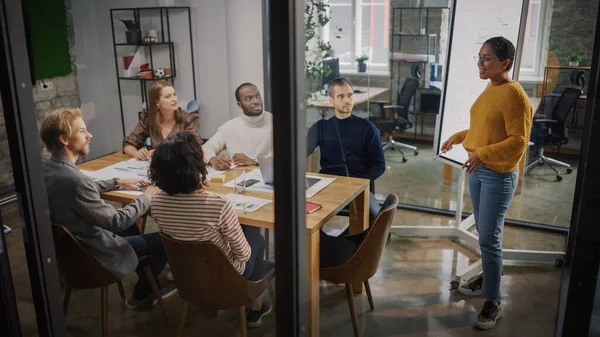 Project Manager Makes a Presentation for a Young Diverse Creative Team in Meeting Room in an Agency. Colleagues Sit Behind Conference Table and Discuss Business Development, User Interface and Design.