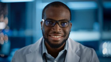 Gözlüklü yakışıklı siyah adam kameraya gülümsüyor ve gülümsüyor. Laboratuvarda çalışan genç zeki erkek mühendis ya da bilim adamı. Teknolojik Bokeh Mavi Arkaplan. Yakın çekim.