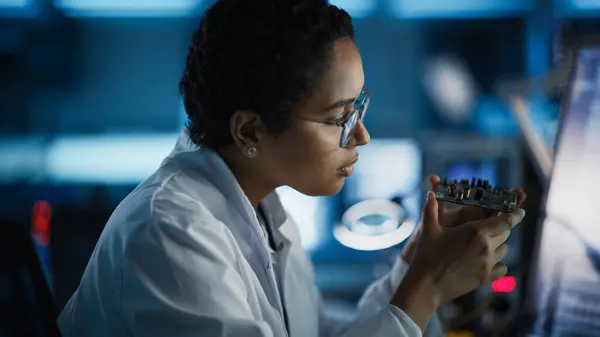 Modern Electronics Research and Development Facility: Beautiful Black Female Engineer Inspects Printed Circuit Board Motherboard. Scientist Designs Industrial PCB, Silicon Microchips, Semiconductors
