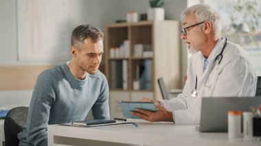 Deneyimli Ortaçağ Aile Doktoru Bir Sağlık Kliniğinde Erkek Hastaya Tablet Bilgisayarda Analiz Sonuçları Gösteriyor. Hastane ofisinde masanın arkasında oturan hekim..