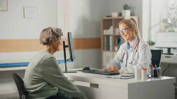 Female Family Doctor is Reading Medical History of Senior Patient and Speaking with Her During Consultation in a Health Clinic. Physician in Lab Coat Sitting Behind a Computer Desk in Hospital Office.