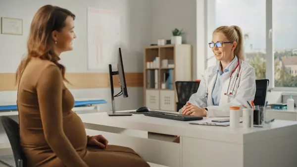 Female Family Doctor is Talking with Young Pregnant Patient During Consultation in a Health Clinic. Experienced Physician in Lab Coat Sitting Behind a Computer Desk in Hospital Office.
