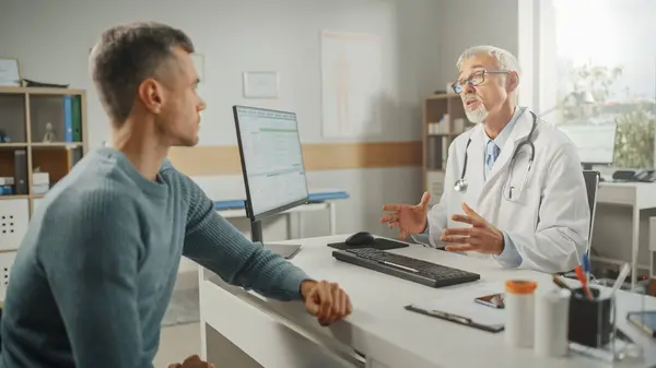 中年家族の医師は 健康クリニックでの相談中に若い男性患者と話しています 病院のパソコン机の後ろに座っている研究室のコートのシニア医師 — ストック写真