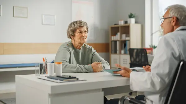 Middle Aged Family Doctor is Talking with Senior Female Patient During Consultation in a Health Clinic. Experienced Caucasian Physician in Lab Coat Sitting Behind a Computer Desk in Hospital Office.