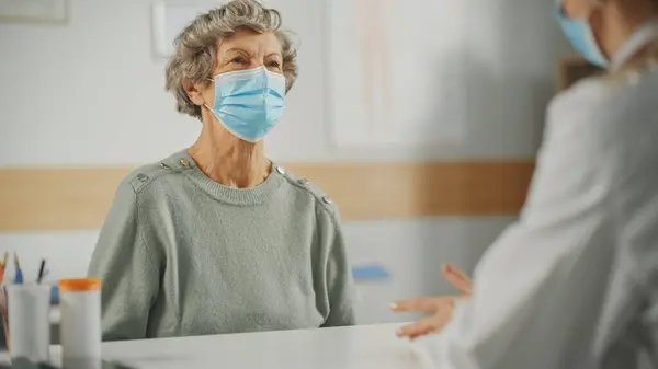 Female Family Doctor is Speaking to a Senior Woman During Consultation in a Health Clinic. Both are Wearing Face Masks. Physician in Lab Coat Prescribing Drugs to Elderly Patient in Hospital Office.