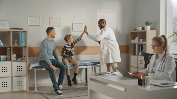 Father Visiting a Friendly Family Doctor with His Young Teenage Son Who Has Broken His Arm, Give High Five. They Talk to an African American Physician in a Hospital Office. Boy is Wearing an Arm Brace
