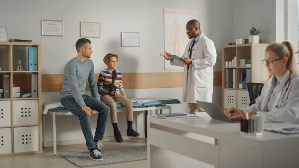 Father Visiting a Friendly Family Doctor with His Young Teenage Son Who Has Broken His Arm. They Talk to an African American Physician in a Hospital Office. Boy is Wearing an Arm Brace.