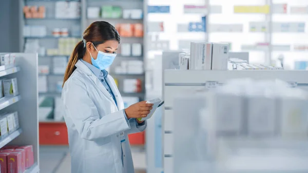 Pharmacy Drugstore: Portrait of Beautiful Pharmacist Wearing Face Mask Uses Digital Tablet Computer, Looks at Camera and Smiles Charmingly, Behind Her Shelves Full of Medicine Packages. Side View Shot