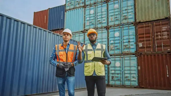Handsome Industrial Engineer and African American Supervisor with Tablet Computer in Hard Hats and Safety Vests Walking in Container Terminal. Colleagues Talk About Logistics Operations.