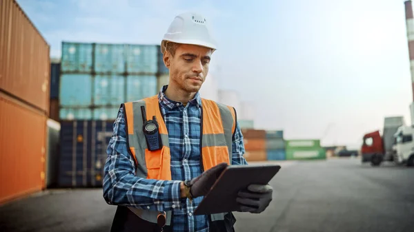Handsome Caucasian Industrial Engineer in White Hard Hat, Orange High-Visibility Vest, Checkered Shirt and Work Gloves Working on Tablet Computer in Shipping Cargo Container Terminal.