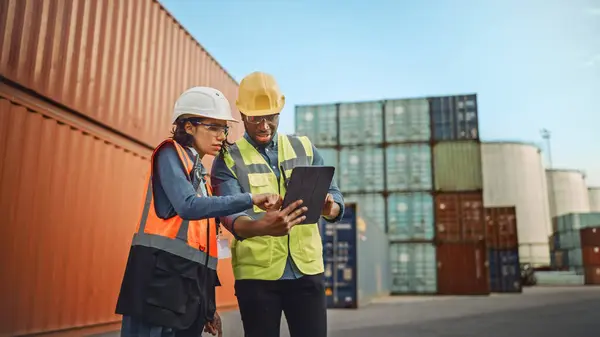Multiethnic Female Industrial Engineer with Tablet and Black African American Male Supervisor in Hard Hats and Safety Vests Stand in Container Terminal. Colleagues Talk About Logistics Operations.