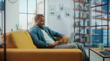 Yakışıklı siyahi adam rahat oturma odasında bir koltukta otururken dizüstü bilgisayarda çalışıyor. Evden çalışan Freelancer. İnternet taraması, Sosyal ağlar kullanımı, Daire 'de eğlenmek.