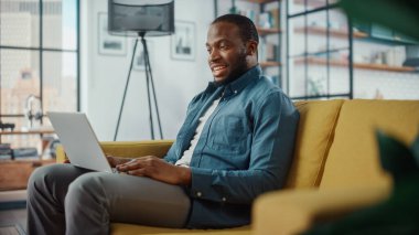Yakışıklı siyahi adam oturma odasında bir koltukta otururken dizüstü bilgisayarında video görüşmesi yapıyor. Evden Çalışan ve İnternet üzerinden İş Arkadaşları ve Müşterilerle Konuşan Serbest Çalışan.