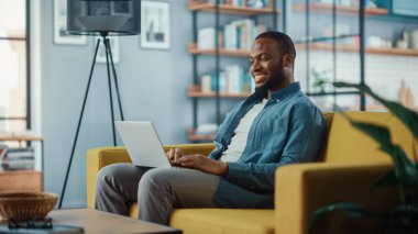 Yakışıklı siyahi adam rahat oturma odasında bir koltukta otururken dizüstü bilgisayarda çalışıyor. Evden çalışan Freelancer. İnternet taraması, Sosyal ağlar kullanımı, Daire 'de eğlenmek.