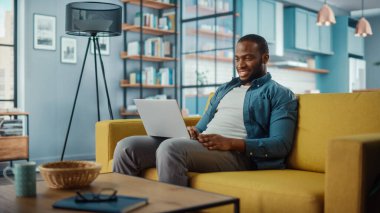 Yakışıklı siyahi adam rahat oturma odasında bir koltukta otururken dizüstü bilgisayarda çalışıyor. Evden çalışan Freelancer. İnternet taraması, Sosyal ağlar kullanımı, Daire 'de eğlenmek.