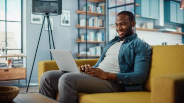 Yakışıklı siyahi adam rahat oturma odasında bir koltukta otururken dizüstü bilgisayarda çalışıyor. Evden çalışan Freelancer. İnternet taraması, Sosyal ağlar kullanımı, Daire 'de eğlenmek.