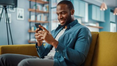 Yakışıklı, Afro-Afrikalı Amerikalı, rahat oturma odasında bir koltukta otururken akıllı telefon kullanıyor. Evden çalışan Freelancer. İnternet taraması, Sosyal ağlar kullanımı, Daire 'de eğlenmek.