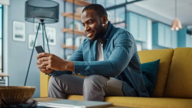 Yakışıklı, Afro-Afrikalı Amerikalı, rahat oturma odasında bir koltukta otururken akıllı telefon kullanıyor. Evden çalışan Freelancer. İnternet taraması, Sosyal ağlar kullanımı, Daire 'de eğlenmek.