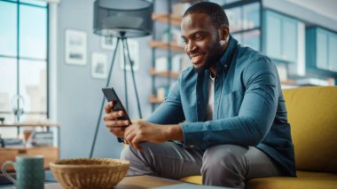 Yakışıklı, Afro-Afrikalı Amerikalı, rahat oturma odasında bir koltukta otururken akıllı telefon kullanıyor. Evden çalışan Freelancer. İnternet taraması, Sosyal ağlar kullanımı, Daire 'de eğlenmek.