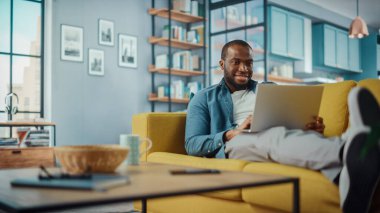Yakışıklı siyahi adam rahat oturma odasında bir koltukta yatarken dizüstü bilgisayarda çalışıyor. Evden çalışan Freelancer. İnternet taraması, Sosyal ağlar kullanımı, Daire 'de eğlenmek.