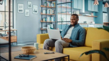 Yakışıklı siyahi adam rahat oturma odasında bir koltukta otururken dizüstü bilgisayarda çalışıyor. Evden çalışan Freelancer. İnternet taraması, Sosyal ağlar kullanımı, Daire 'de eğlenmek.