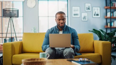 Yakışıklı siyahi adam rahat oturma odasında bir koltukta otururken dizüstü bilgisayarda çalışıyor. Evden çalışan Freelancer. İnternet taraması, Sosyal ağlar kullanımı, Daire 'de eğlenmek.
