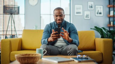Oturma odasında koltukta dinlenirken akıllı telefon kullanan heyecanlı siyahi Amerikalı adam. Mutlu Adam Evde Gülümsüyor ve İnternet üzerinden İş Arkadaşları ve Müşterilerle Sohbet Ediyor. Sosyal ağlar kullanılıyor.