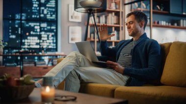 Yakışıklı beyaz adam rahat oturma odasında bir koltukta otururken dizüstü bilgisayarında video görüşmesi yapıyor. Evden Çalışan ve İnternet üzerinden İş Arkadaşları ve Müşterilerle Konuşan Serbest Çalışan.