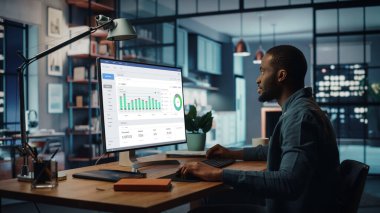 Yaratıcı Ev Oturma Odasında Masaüstü Bilgisayarı 'nda çalışan yakışıklı siyahi Amerikalı Uzman. Serbest Erkek, Pazar Analizi Yapıyor ve Müşteriler ve İşverenler İçin Çizelge Oluşturuyor.