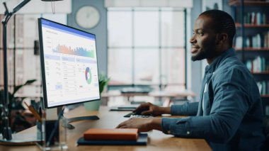 Yaratıcı Ev Oturma Odasında Masaüstü Bilgisayarı 'nda çalışan yakışıklı siyahi Amerikalı Uzman. Serbest Erkek, Pazar Analizi Yapıyor ve Müşteriler ve İşverenler İçin Çizelge Oluşturuyor.