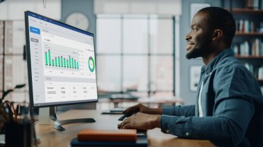 Yaratıcı Ev Oturma Odasında Masaüstü Bilgisayarı 'nda çalışan yakışıklı siyahi Amerikalı Uzman. Serbest Erkek, Pazar Analizi Yapıyor ve Müşteriler ve İşverenler İçin Çizelge Oluşturuyor.