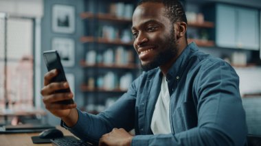 Oturma odasında Masa 'da otururken akıllı telefon kullanan Afro-Amerikan Adam' ın kutlu olsun. Heyecanlı Adam Evde Gülümsüyor ve İnternet Üzerindeki Meslektaşlar, Müşteriler ve Arkadaşlarla Konuşuyor.