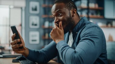 Oturma odasında otururken akıllı telefon kullanan siyahi Afrikalı Amerikalı adam şok oldu. İnsan, İnternet ya da Sosyal ağlar üzerinden Aile Üyeleri ve Arkadaşlarıyla Konuşurken Korkunç Haberler Aldı.