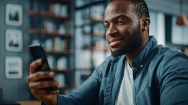 Oturma odasında Masa 'da otururken akıllı telefon kullanan Afro-Amerikan Adam' ın kutlu olsun. Heyecanlı Adam Evde Gülümsüyor ve İnternet Üzerindeki Meslektaşlar, Müşteriler ve Arkadaşlarla Konuşuyor.