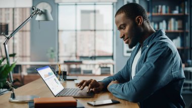 Yaratıcı Ev Oturma Odasında dizüstü bilgisayar üzerinde çalışan yakışıklı siyahi Amerikalı Uzman. Serbest Erkek, Pazar Analizi Yapıyor ve Müşteriler ve İşverenler İçin Çizelge Oluşturuyor.