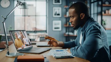 Yakışıklı Afrikalı Amerikalı Adam Oturma Odasında Masa başında otururken dizüstü bilgisayarında video görüşmesi yapıyor. Evden Çalışan ve İnternet üzerinden İş Arkadaşları ve Müşterilerle Konuşan Serbest Çalışan.